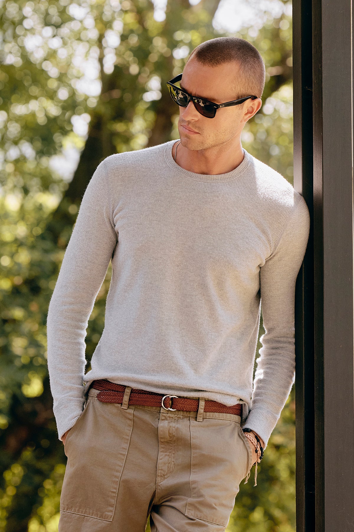 A man in sunglasses and a light sweater leans against a post, perfectly showcasing the BECKER CREW by Velvet by Graham & Spencer. The fitted crew neck design enhances his style as he stands outdoors, with trees providing a picturesque backdrop.-38426511474881