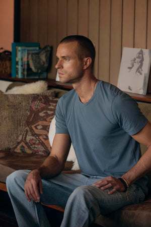 A man with short hair sits on a couch, clad in the HOWARD TEE by Velvet by Graham & Spencer, made from lightweight Pima cotton, paired with jeans. He gazes to the side with a focused expression. The background features a vintage atmosphere, complete with a wall adorned by books and framed artwork.