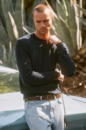 A man in a MARCO SWEATER by Velvet by Graham & Spencer, characterized by its cotton cashmere blend and seed stitch texture, paired with light jeans is standing outdoors, leaning against a light blue car. He has a serious expression and is touching his chin with one hand.