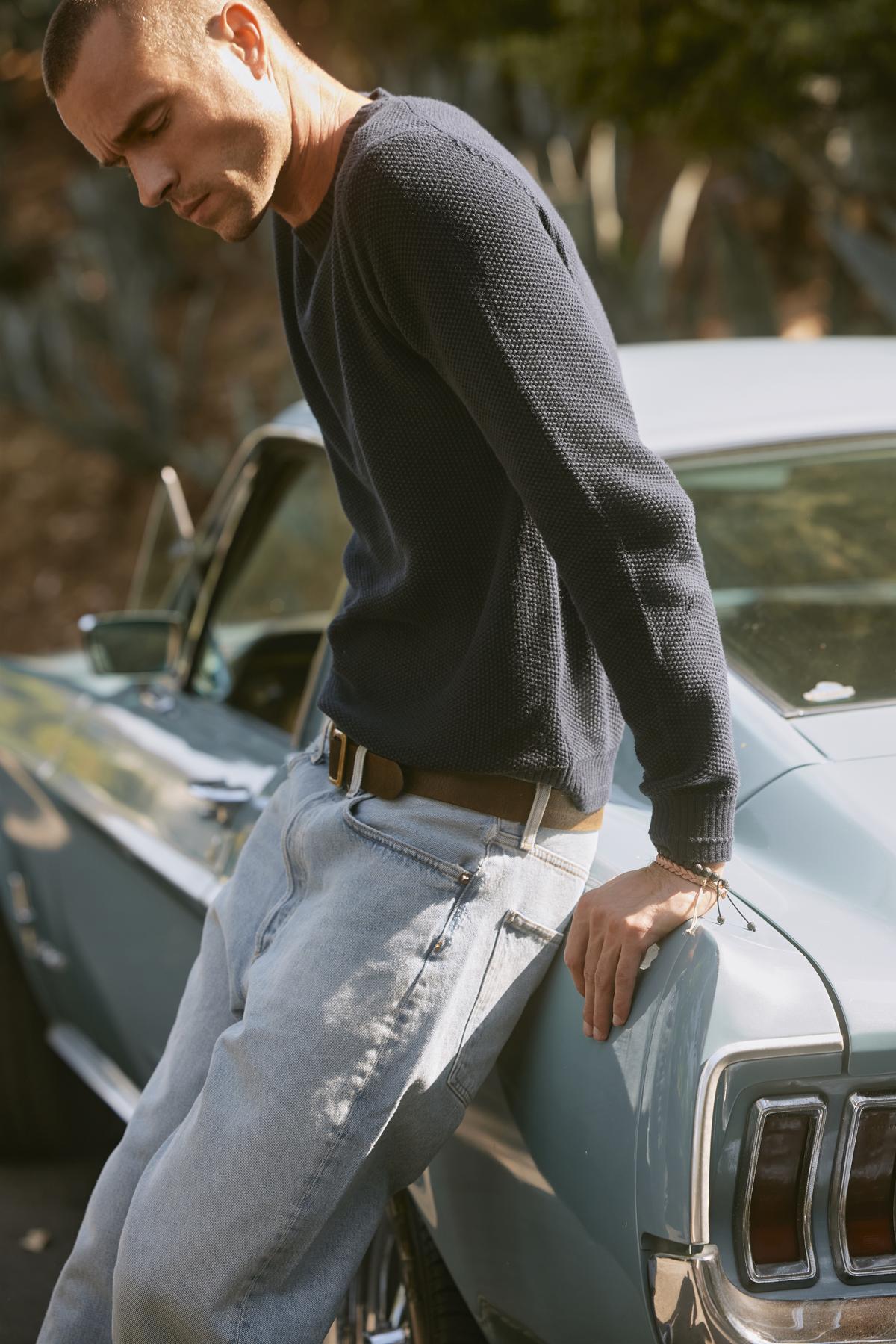   A man in a MARCO SWEATER by Velvet by Graham & Spencer and light jeans leans against an old-fashioned blue car, looking down. 