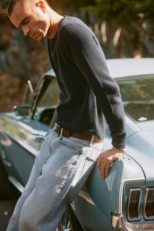 A man in a MARCO SWEATER by Velvet by Graham & Spencer and light jeans leans against an old-fashioned blue car, looking down.