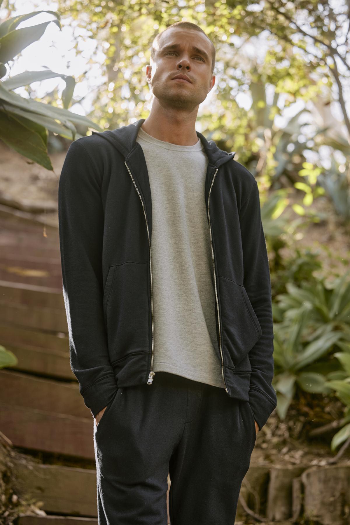   A man stands outdoors in casual attire, sporting the RODAN HOODIE by Velvet by Graham & Spencer along with pants and a light grey t-shirt underneath. He is looking upwards, surrounded by greenery and steps in the background, making his hoodie a perfect workout layer for the serene setting. 