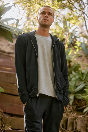 A man stands outdoors in casual attire, sporting the RODAN HOODIE by Velvet by Graham & Spencer along with pants and a light grey t-shirt underneath. He is looking upwards, surrounded by greenery and steps in the background, making his hoodie a perfect workout layer for the serene setting.