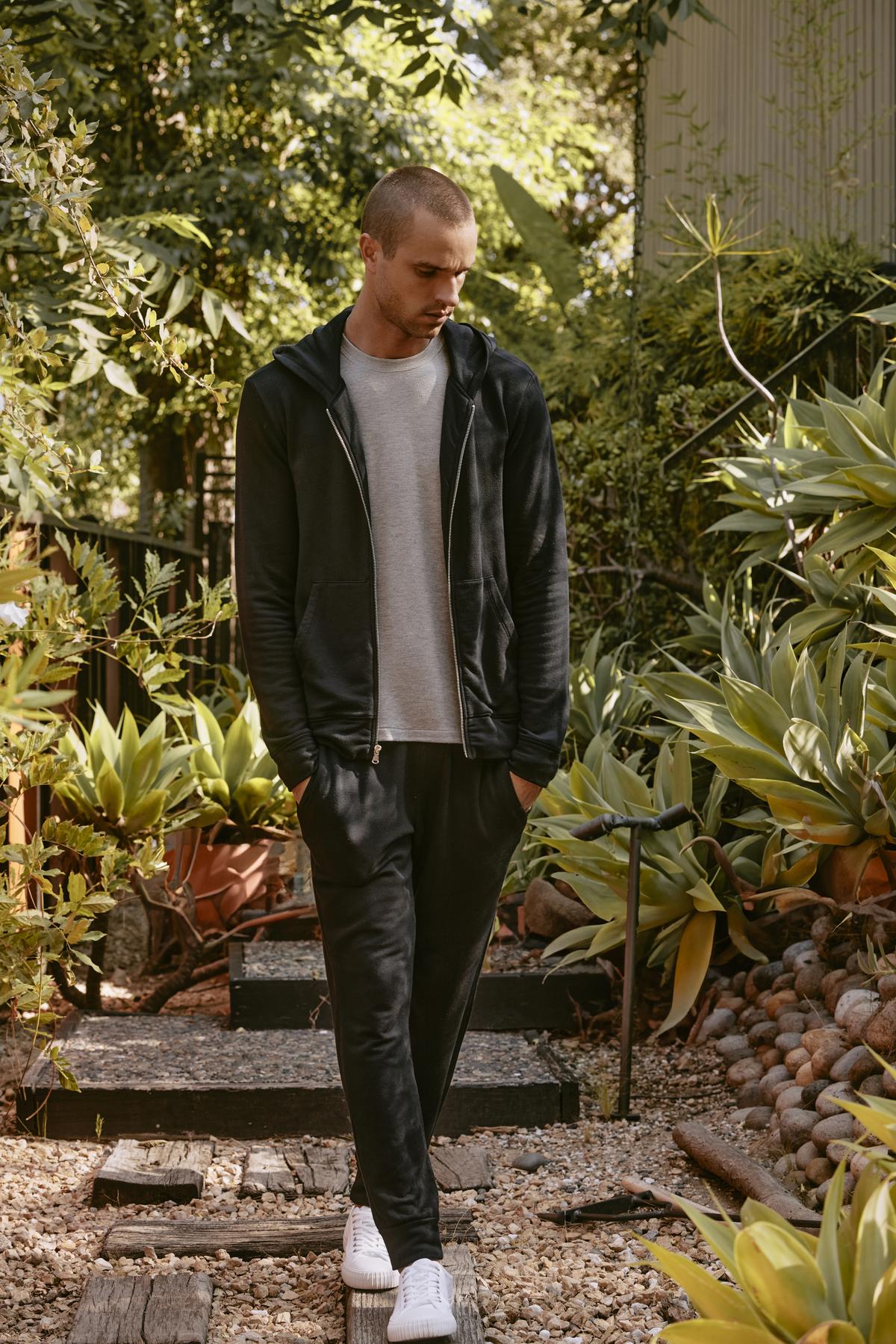   A man wearing a Velvet by Graham & Spencer RODAN HOODIE, styled with a non-bulky fit, along with a gray t-shirt, black pants, and white sneakers walks down a garden path lined with greenery on a sunny day. 