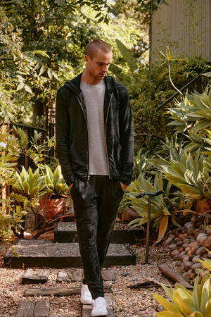 A man wearing a Velvet by Graham & Spencer RODAN HOODIE, styled with a non-bulky fit, along with a gray t-shirt, black pants, and white sneakers walks down a garden path lined with greenery on a sunny day.