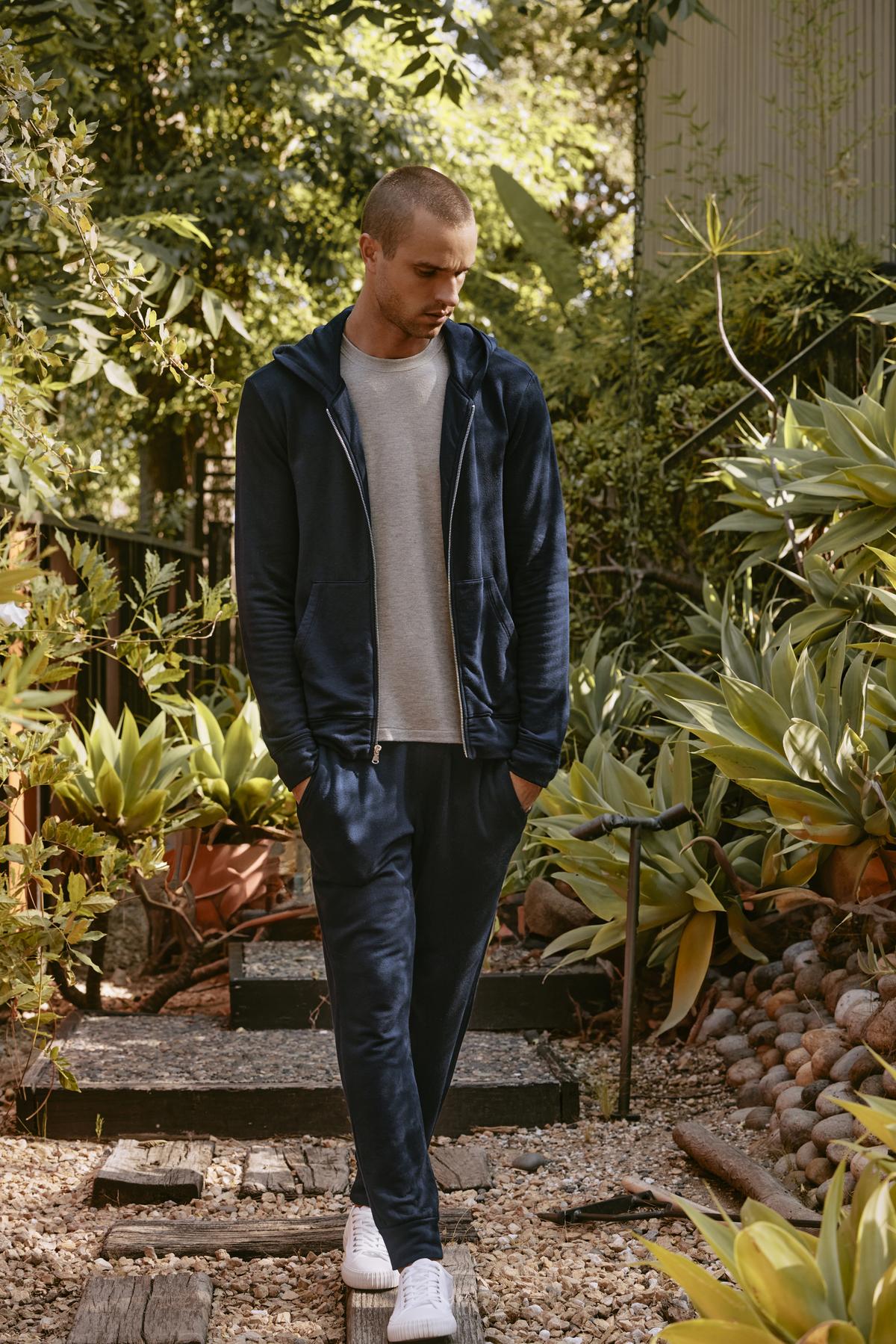 A person in a tracksuit featuring Velvet by Graham & Spencer's CROSBY JOGGER and white sneakers stands on a garden path surrounded by lush green plants.-38457202114753