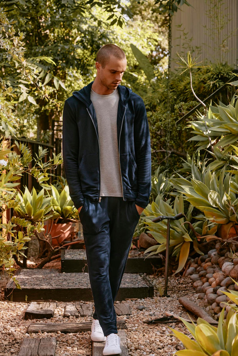 A person in a tracksuit featuring Velvet by Graham & Spencer's CROSBY JOGGER and white sneakers stands on a garden path surrounded by lush green plants.