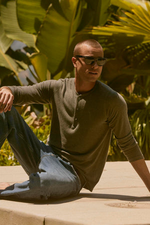 A man in the stylish RYLAND HENLEY by Velvet by Graham & Spencer sits outdoors on a sunny day, wearing sunglasses with large green leaves in the background, embodying the perfect style for casual occasions.