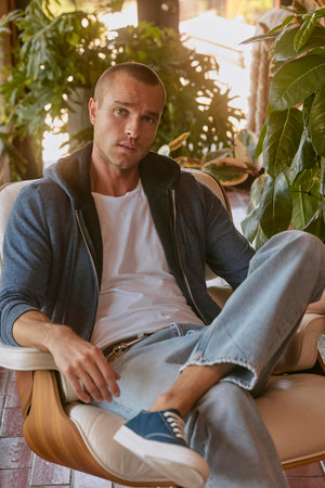 A man with short hair in a white t-shirt, Velvet by Graham & Spencer's SALVADORE HOODIE, and jeans sits in a chair indoors, surrounded by green plants.