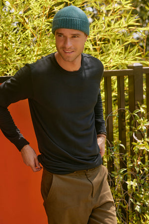 A person wearing a green beanie and the SOREN PULLOVER, a black long-sleeve shirt made of medium-weight extra soft luxe fleece from Velvet by Graham & Spencer, stands outdoors in front of a wooden fence with greenery in the background.