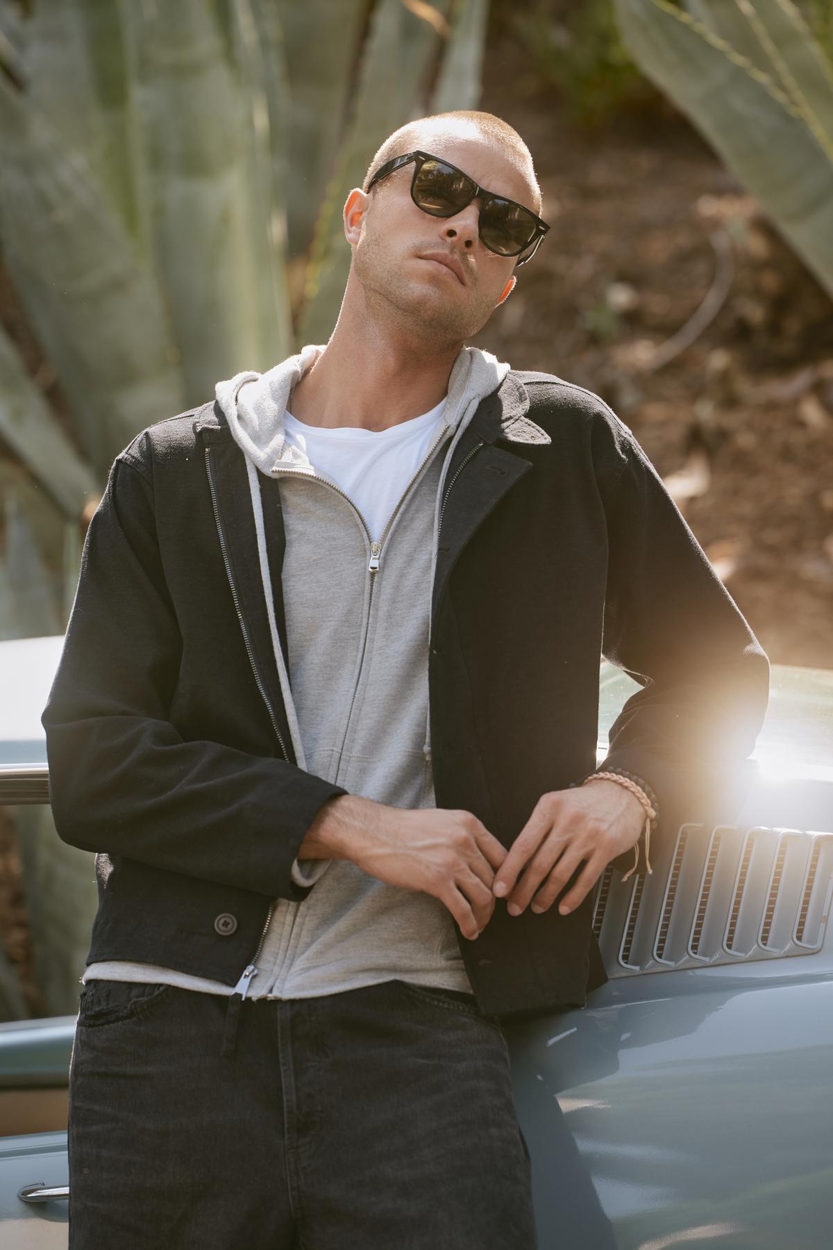 A person in sunglasses leans against a vintage car outdoors, sporting the WILBUR JACKET by Velvet by Graham & Spencer, crafted from a brushed cotton blend over a gray hoodie, with plants in the background.-38247288144065