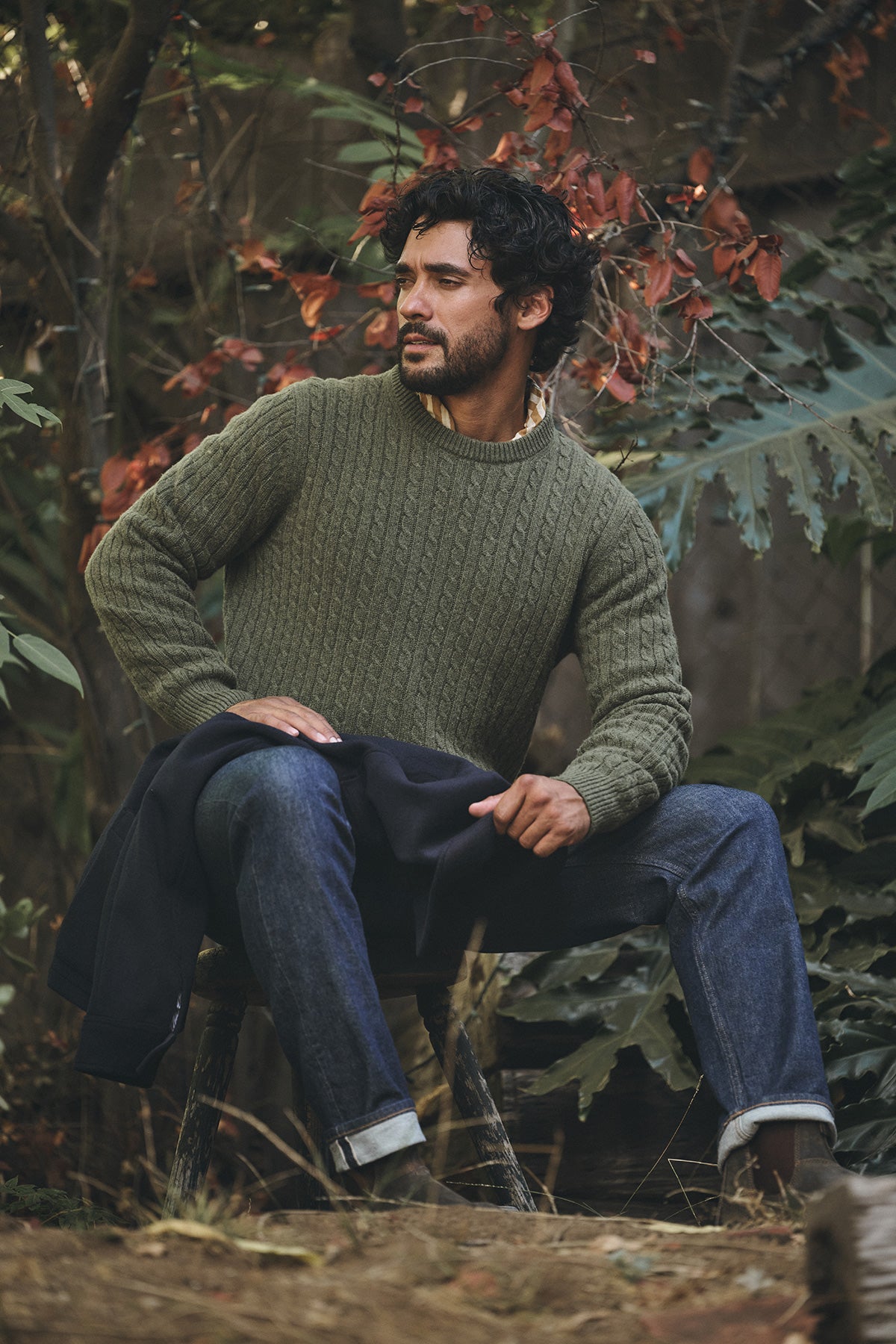 A man sits on a wooden stool outdoors among autumn leaves, wearing the ALONZO SWEATER by Velvet by Graham & Spencer and jeans.-40134784090305