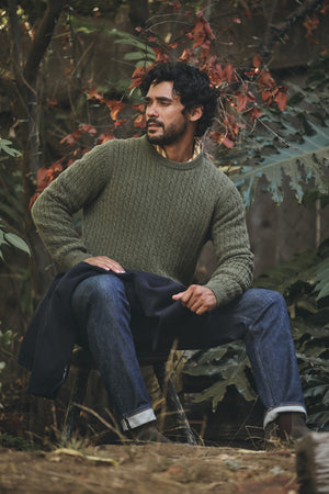 A man sits on a wooden stool outdoors among autumn leaves, wearing the ALONZO SWEATER by Velvet by Graham & Spencer and jeans.