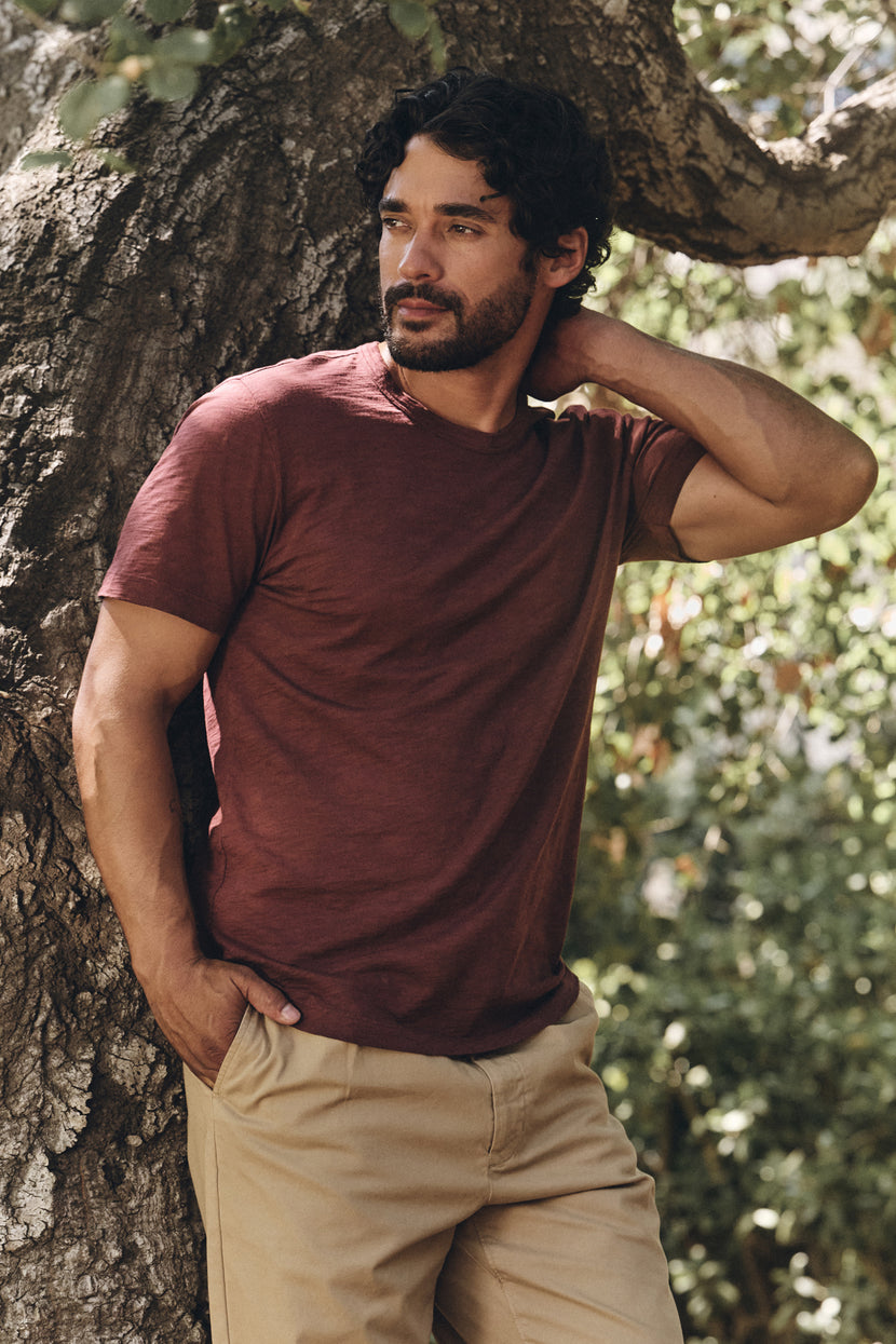 A man stands outdoors by a tree, wearing Velvet by Graham & Spencer's AMARO TEE in maroon Peruvian cotton with a crew neckline and beige pants, one hand in his pocket and the other touching his neck.