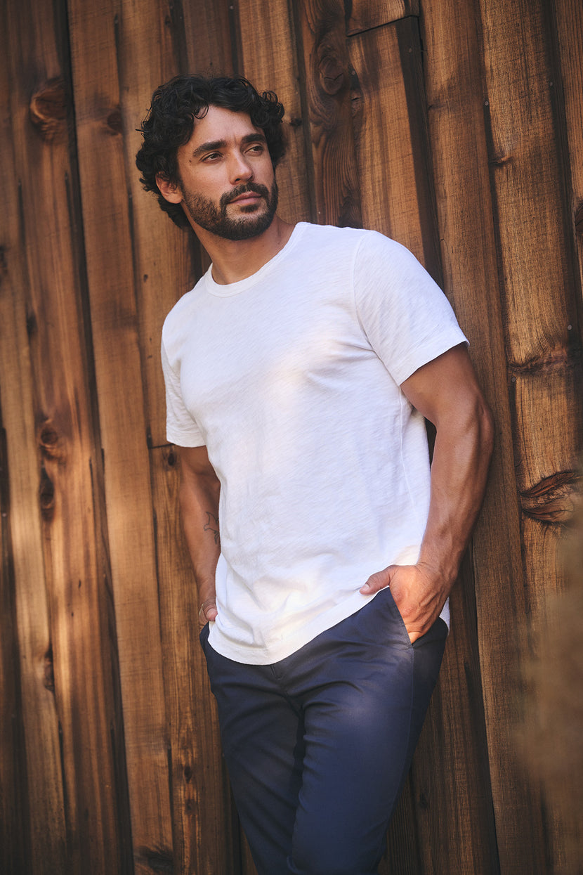 A man with curly hair and a beard stands against a wooden wall, wearing the AMARO TEE by Velvet by Graham & Spencer—a Peruvian cotton white t-shirt with a classic crew neckline—making it an ideal layering piece for any occasion.