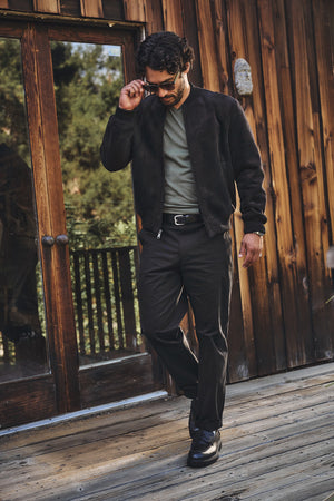 A man in sunglasses models the Velvet by Graham & Spencer AMIAS BOMBER JACKET—a faux suede bomber with sherpa lining—paired with a green t-shirt, black pants, and black shoes on a wooden deck before a rustic building.