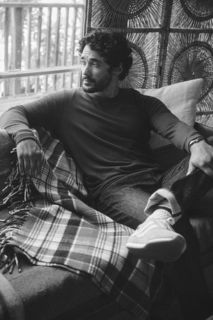 A man in the BECKER CREW by Velvet by Graham & Spencer sits on a sofa with a plaid blanket, gazing out the window. He pairs this cozy jersey with jeans and sneakers in a black and white scene of calm reflection.