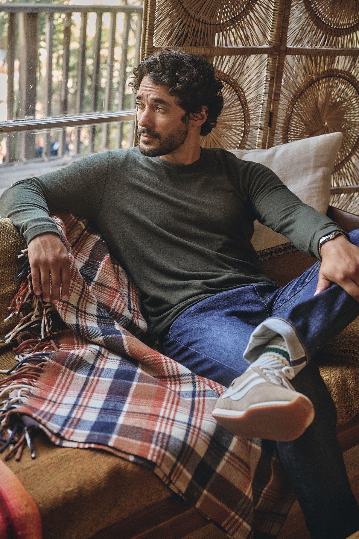 A man with curly hair sits on a couch with a plaid blanket, wearing the BECKER CREW by Velvet by Graham & Spencer—a cozy dark green long-sleeve top—paired with blue jeans and sneakers as he looks to the side.-40247441785025