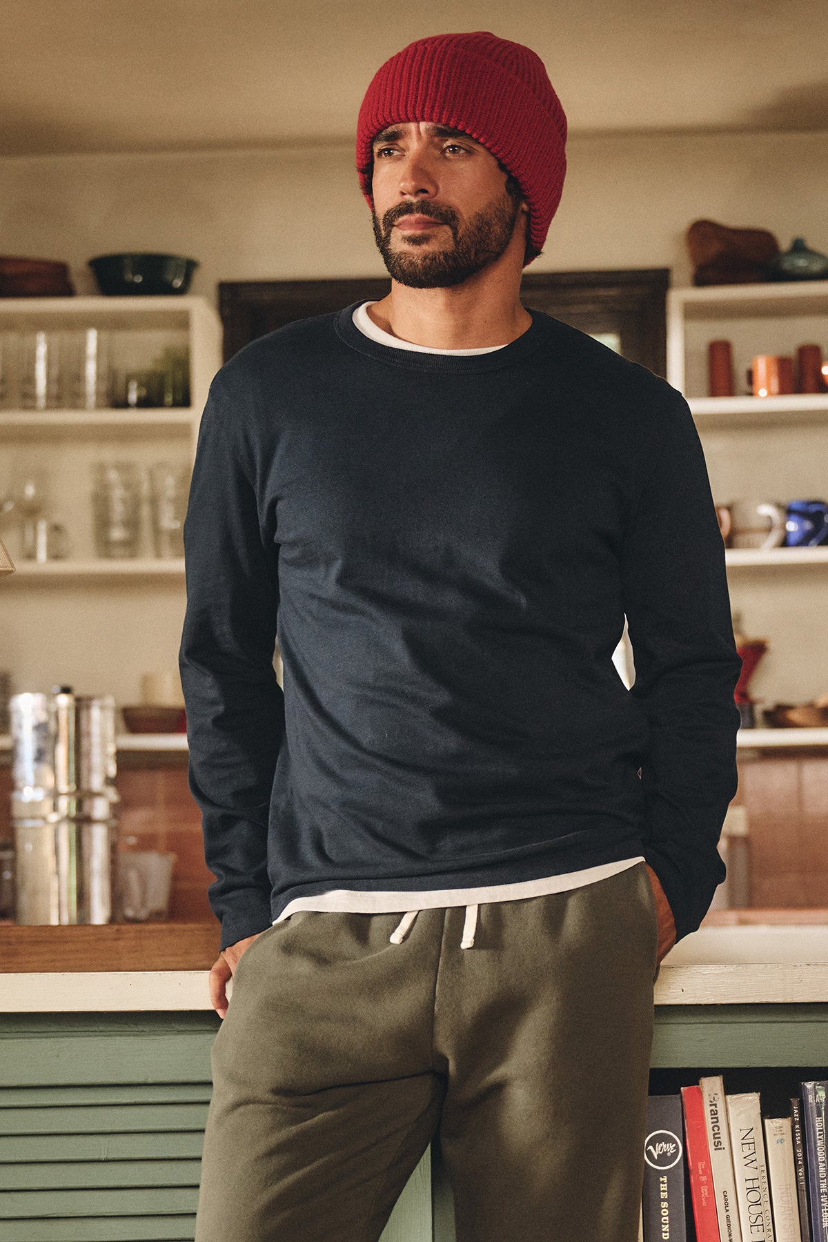 A man with a beard, in casual wear—a Velvet by Graham & Spencer BRADY TEE, red knit beanie, and olive sweatpants—leans against a kitchen counter in a cozy home.-40277087355073