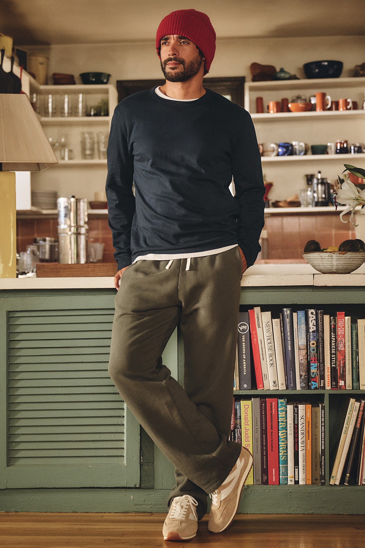 A man in casual attire—a red knit beanie, navy long-sleeve shirt, and the HUDSON SWEATPANT by Velvet by Graham & Spencer—stands by a kitchen counter with books and dishware in the background.-40277063401665