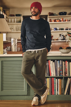 A man in casual attire—a red knit beanie, navy long-sleeve shirt, and the HUDSON SWEATPANT by Velvet by Graham & Spencer—stands by a kitchen counter with books and dishware in the background.