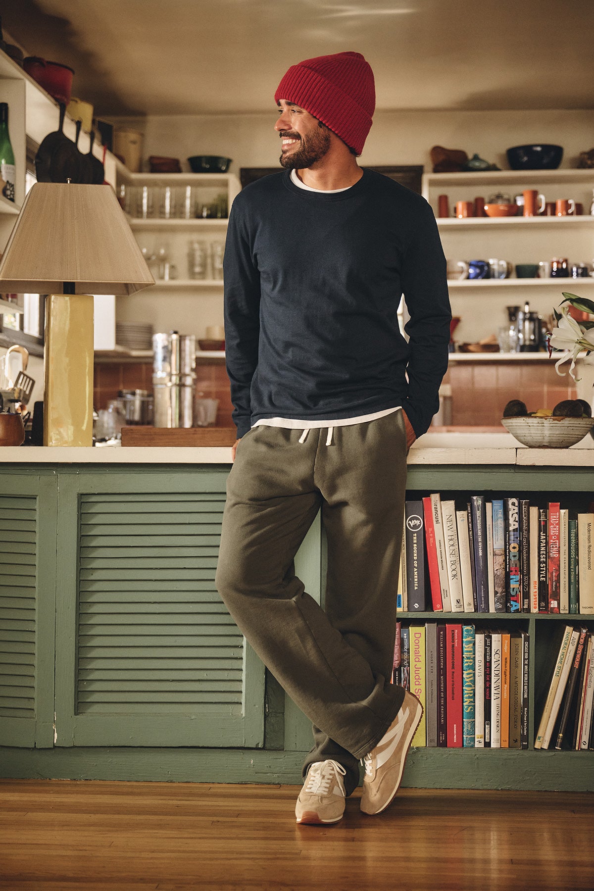 A man wearing the Velvet by Graham & Spencer BRADY TEE, a midweight dark long-sleeve jersey, paired with a red beanie and olive joggers, stands leaning against a kitchen counter with bookshelves, looking to the side and smiling.-40277087322305