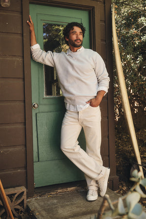 A man wearing the Velvet by Graham & Spencer CILLIAN SWEATSHIRT, white pants, and white sneakers stands by a green door, leaning casually against the doorframe with one hand in his pocket.