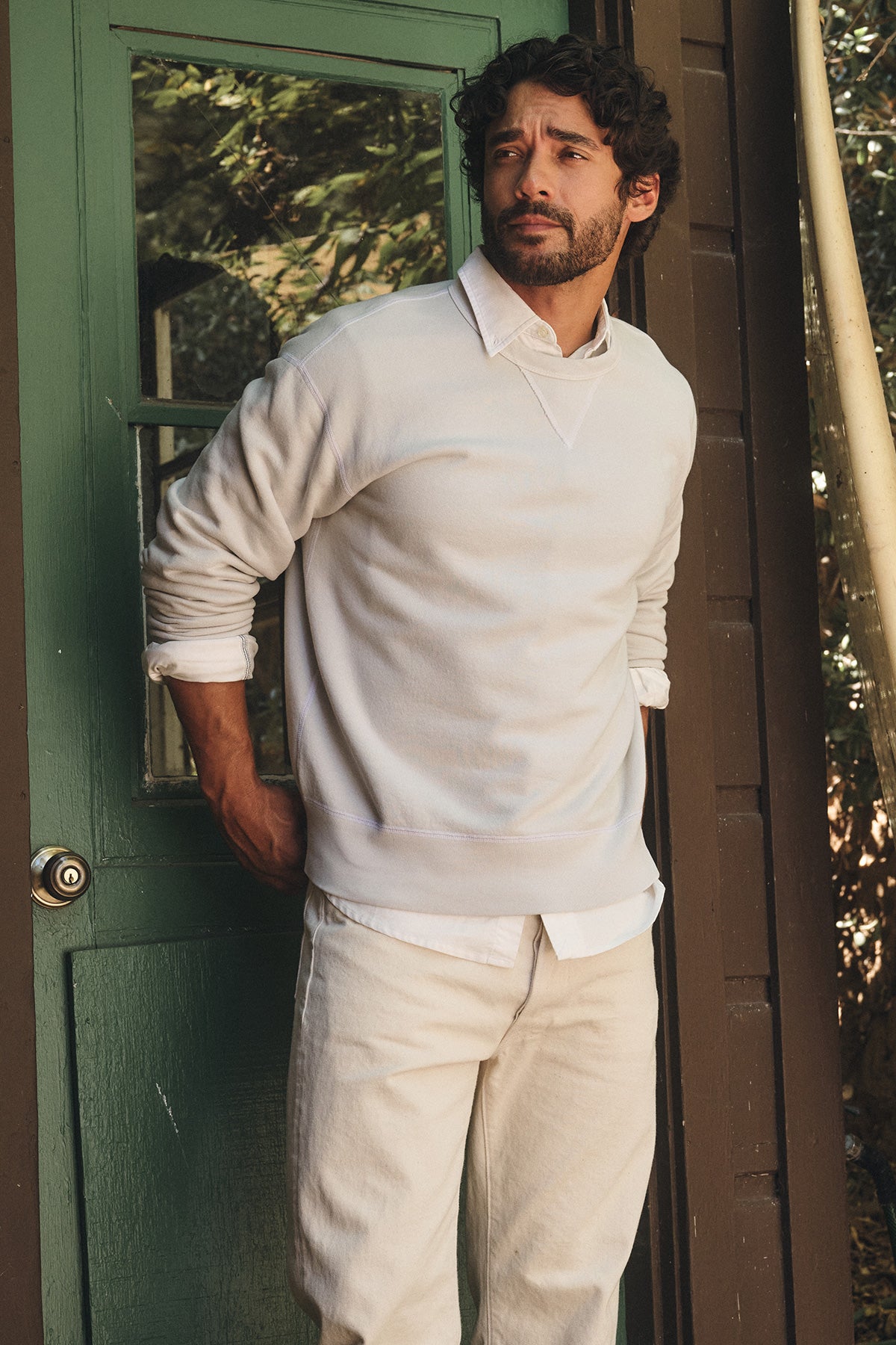 A man looks to the side, wearing the CILLIAN SWEATSHIRT by Velvet by Graham & Spencer over a white collared shirt and light pants, standing in front of a green door.