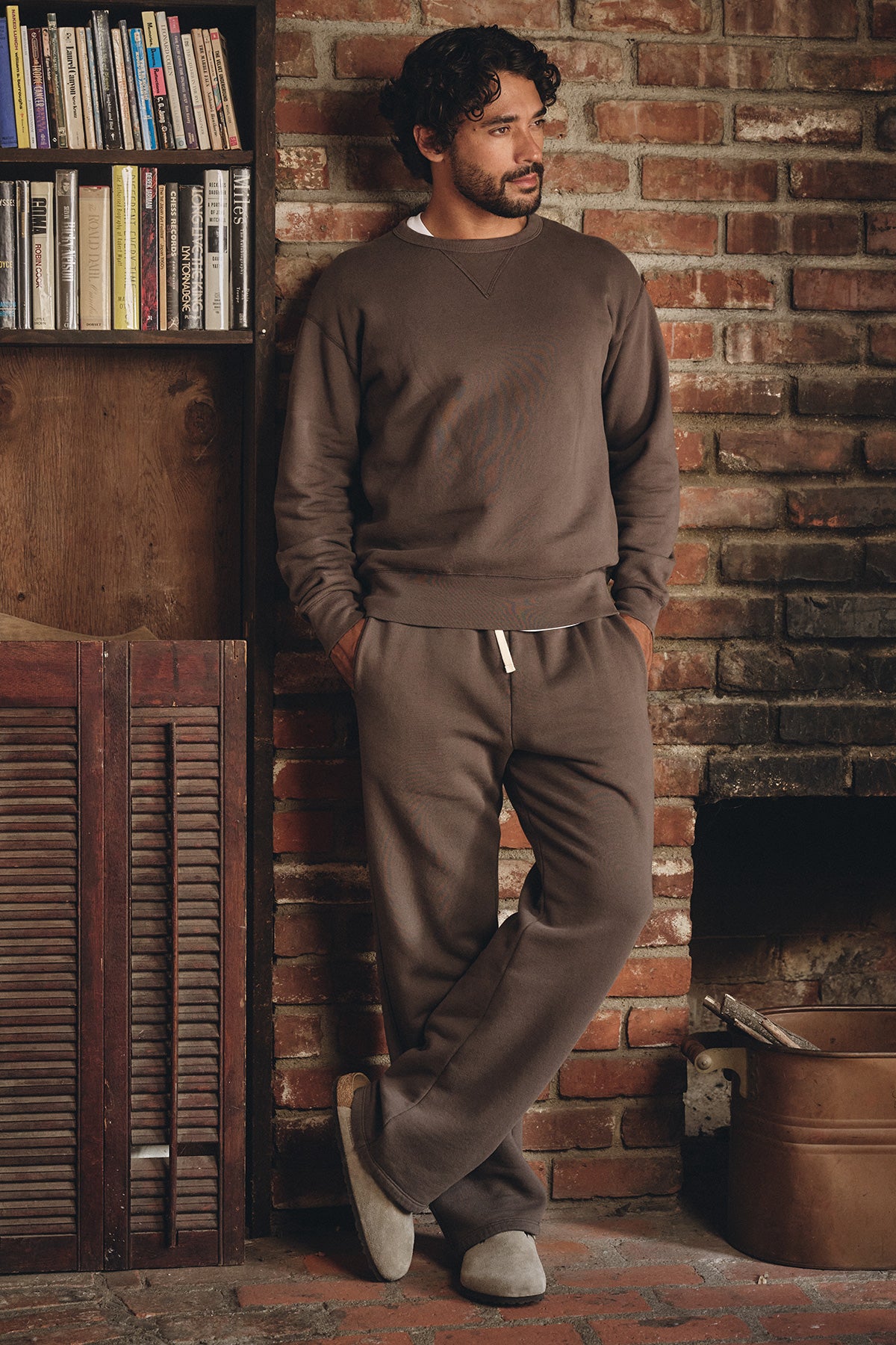 A man wears Velvet by Graham & Spencer's HUDSON SWEATPANT, standing by a brick wall near a bookshelf and fireplace, hands in pockets—the perfect choice for effortless everyday comfort.