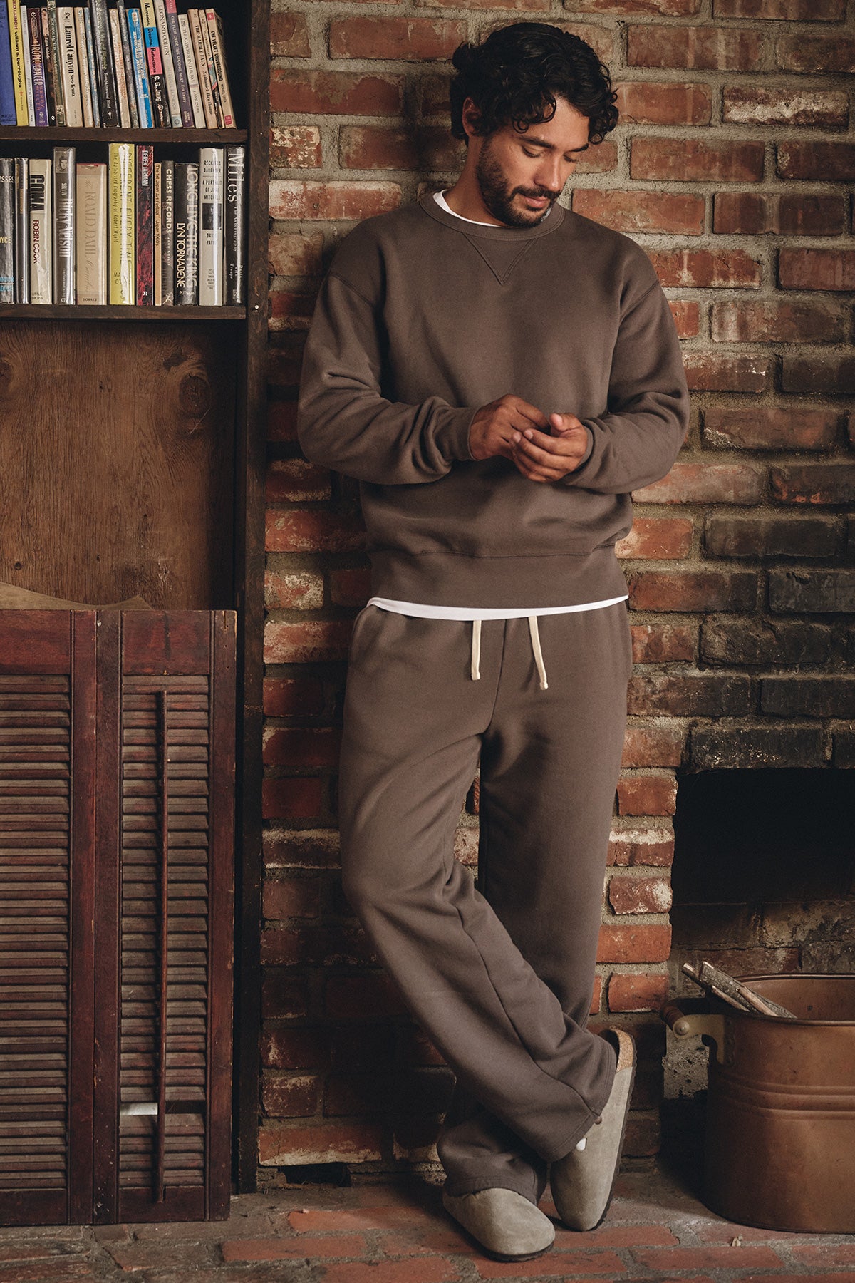 A man wearing the CILLIAN SWEATSHIRT by Velvet by Graham & Spencer stands by a brick wall, looking at his phone near a bookshelf and fireplace.