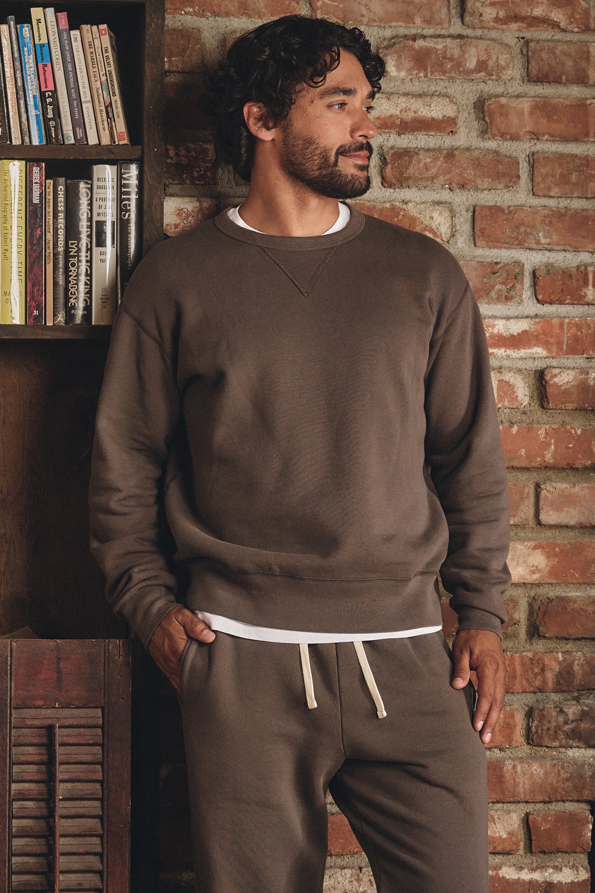 A man stands indoors by a brick wall, wearing the CILLIAN SWEATSHIRT by Velvet by Graham & Spencer and matching sweatpants over a white t-shirt, with one hand in his pocket.-40277059272897