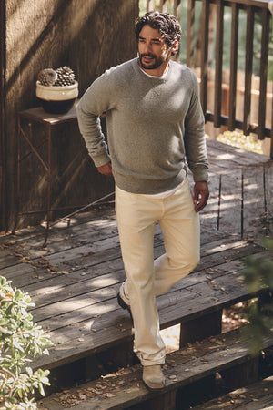 A man stands on a wooden deck wearing the Velvet by Graham & Spencer CORTEZ SWEATER, styled with a white shirt, cream pants, and beige shoes; a small table and potted plants appear in the background.