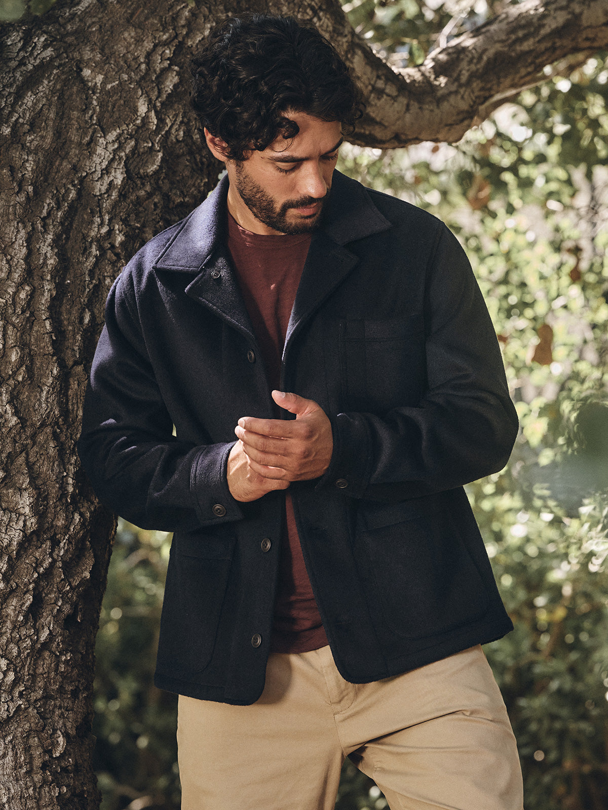 A man with curly hair stands by a tree in the EMERSON CHORE JACKET by Velvet by Graham & Spencer, paired with a maroon shirt and beige pants—an understated style ideal for everyday wear.