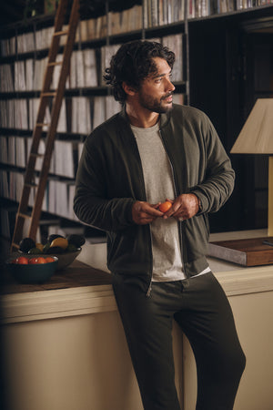 A man wearing the MILES JACKET by Velvet by Graham & Spencer stands at a kitchen counter holding two tomatoes, with a bowl of assorted fruit and shelves of records in the background.