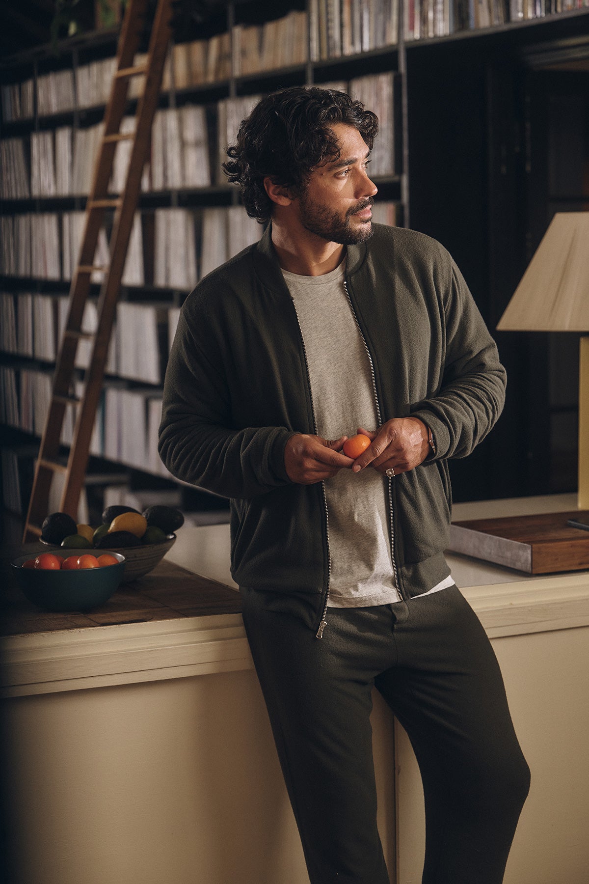 Wearing ZEALAND SWEATPANT by Velvet by Graham & Spencer, a man with an elasticized drawstring waist lounges by a counter with tomatoes, near fruit and recordshelves, and a ladder in the background.