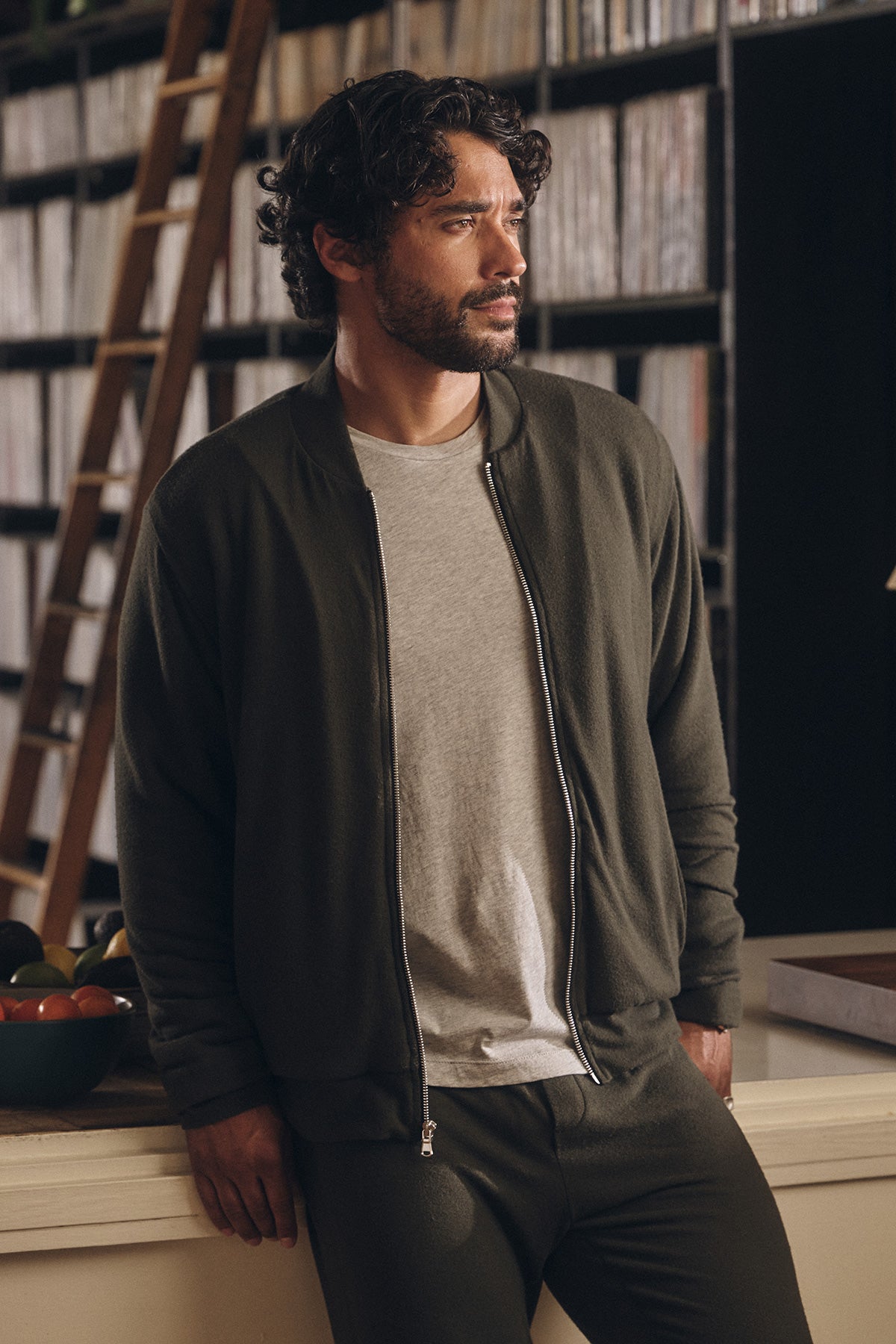 A man wearing the MILES JACKET by Velvet by Graham & Spencer stands indoors near a counter, with shelves of books or records and a ladder visible in the background.