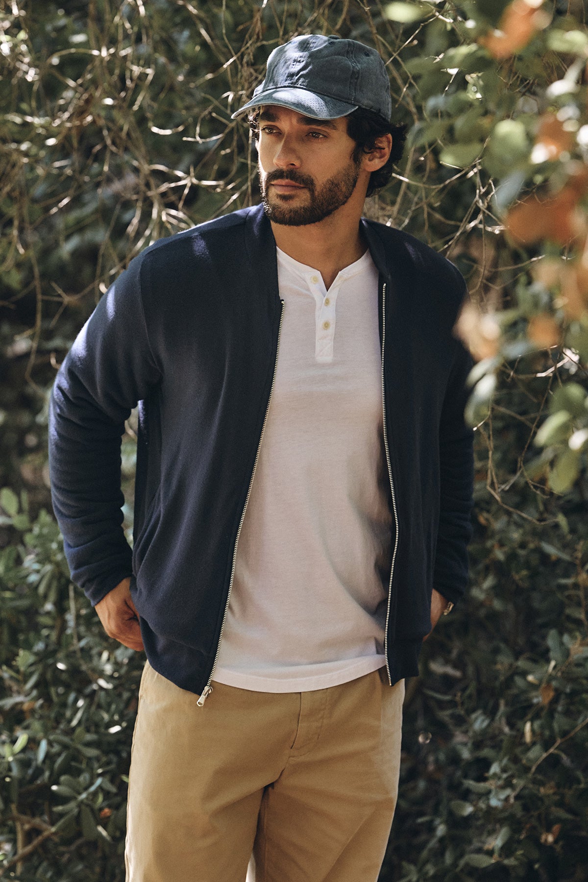 A man in the Velvet by Graham & Spencer MILES JACKET, a soft knit-shell casual jacket, paired with a white henley and khaki pants, stands outdoors near leafy branches.-40134889210049