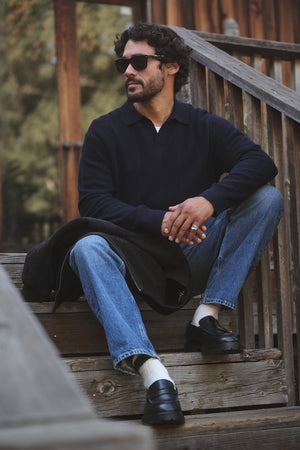 A man in sunglasses, blue jeans, black loafers, and the Velvet by Graham & Spencer REDDING SWEATER POLO sits on outdoor wooden steps with a cigarette—demonstrating versatile casual layering.