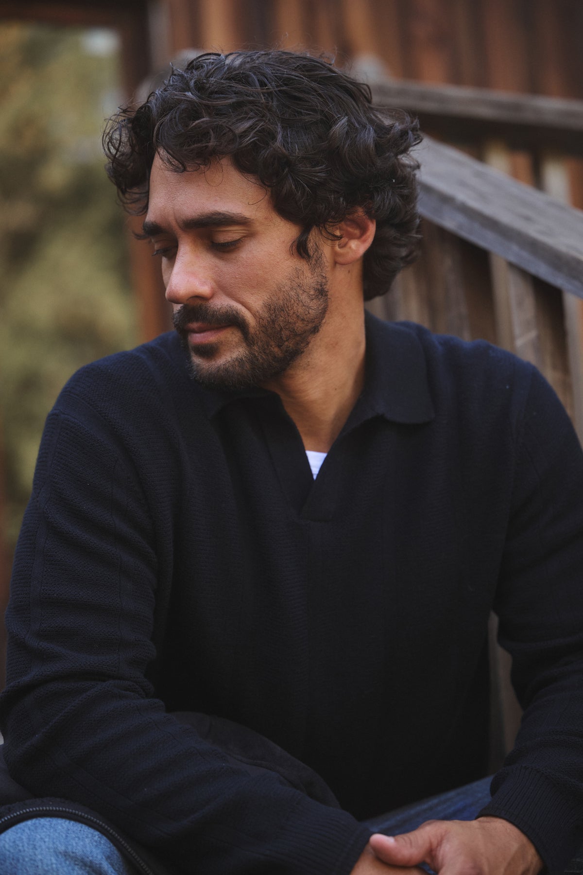 A man with curly dark hair and a beard sits outdoors in front of a wooden railing, looking down neutrally, wearing the REDDING SWEATER POLO by Velvet by Graham & Spencer and jeans.-40054247588033