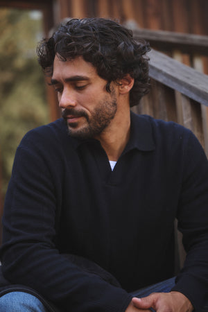 A man with curly dark hair and a beard sits outdoors in front of a wooden railing, looking down neutrally, wearing the REDDING SWEATER POLO by Velvet by Graham & Spencer and jeans.