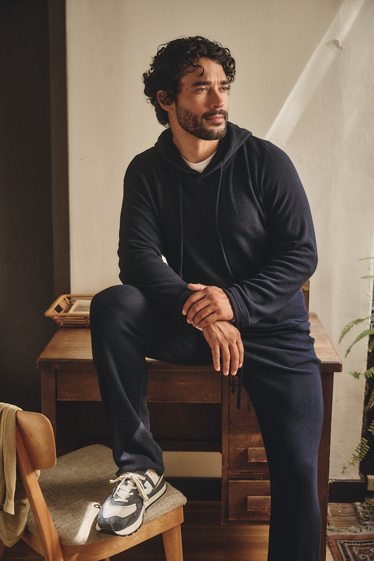 A man with curly hair and a beard sits on a desk’s edge, wearing a dark hooded sweatshirt, Velvet by Graham & Spencer’s OZZY SWEATPANT, and sneakers in a softly lit room.-40134823870657