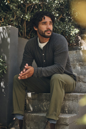 A man with dark hair and a beard sits on outdoor steps in the RYLAND HENLEY by Velvet by Graham & Spencer, paired with olive pants and brown shoes, with greenery in the background.