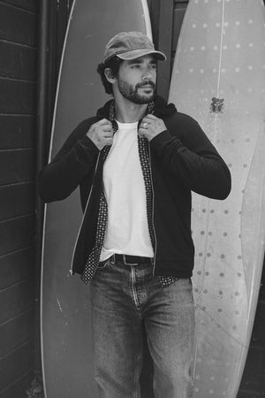 A man in a cap, white t-shirt, and jeans adjusts his Velvet by Graham & Spencer SALVADORE HOODIE while standing in front of two surfboards.