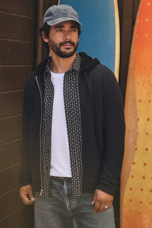 A man with a beard wears a blue cap and the SALVADORE HOODIE by Velvet by Graham & Spencer, featuring faux shearling lining, over a white t-shirt and patterned shirt, standing before two upright surfboards.