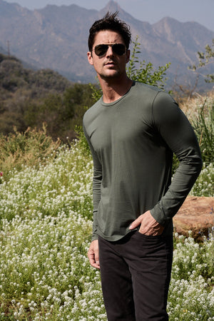Model standing outdoors wearing Skeeter Crew Neck Long Sleeve Tee in olive and sunglasses front