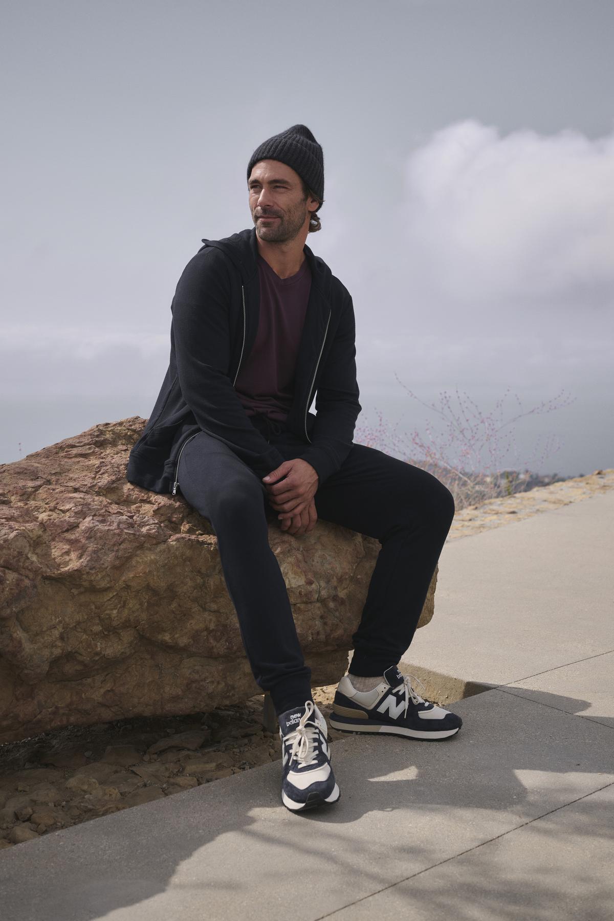   Dressed in casual attire, a person relaxes on a rock outdoors beneath a cloudy sky, wearing the CROSBY JOGGER by Velvet by Graham & Spencer. Their beanie complements the joggers' elasticized drawstring waist, and their sneakers sit comfortably on the rugged terrain. 