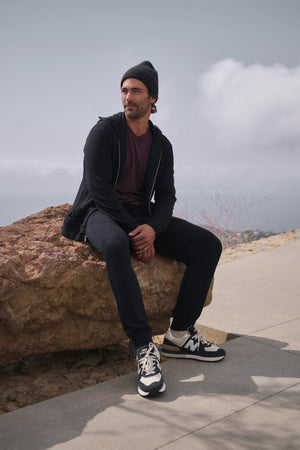 Dressed in casual attire, a person relaxes on a rock outdoors beneath a cloudy sky, wearing the CROSBY JOGGER by Velvet by Graham & Spencer. Their beanie complements the joggers' elasticized drawstring waist, and their sneakers sit comfortably on the rugged terrain.