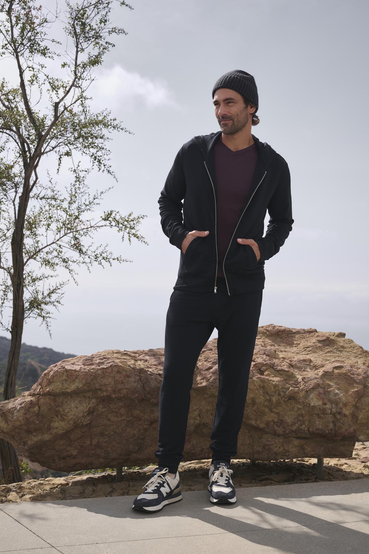   A person stands outdoors wearing a black hoodie, a beanie, and sneakers. Their CROSBY JOGGER by Velvet by Graham & Spencer enhances their casual look with its sleek design as they stand with hands in pockets in front of a large rock and tree. 