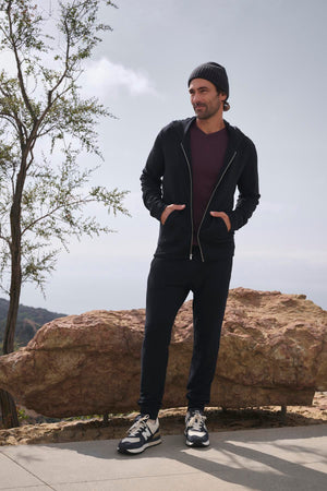 A person stands outdoors wearing a black hoodie, a beanie, and sneakers. Their CROSBY JOGGER by Velvet by Graham & Spencer enhances their casual look with its sleek design as they stand with hands in pockets in front of a large rock and tree.