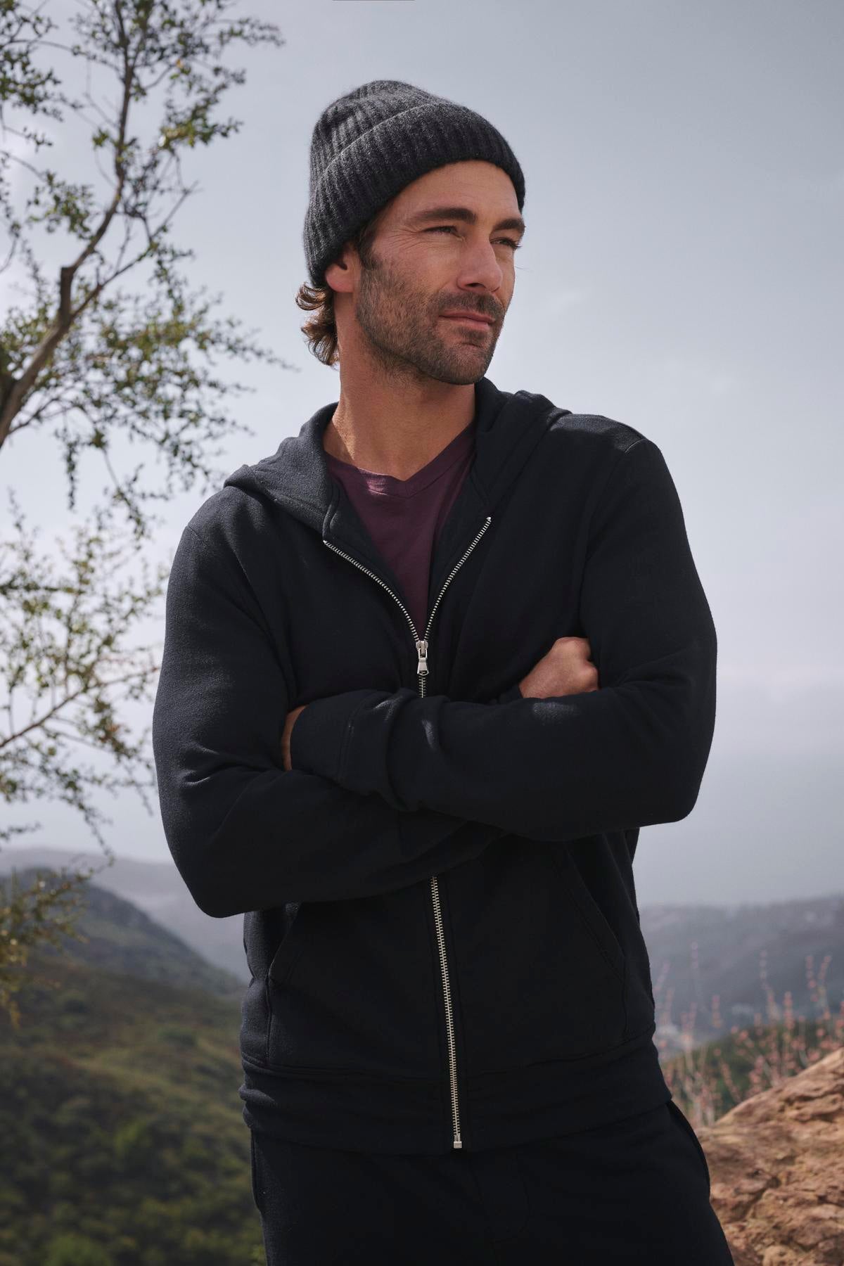 A person is standing outdoors, with arms crossed and facing the camera, wearing the RODAN HOODIE by Velvet by Graham & Spencer. The backdrop consists of trees and hills.-38466938765505